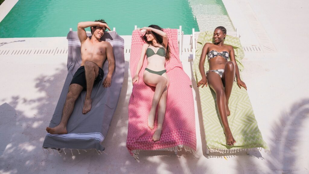 Ein Mann und zwei Frauen entspannen auf ANNA ANIQ Badetüchern auf Poolliegen.