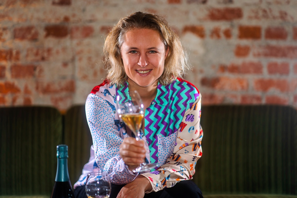 Nicola Neumann, Geschäftsführerin von Champagne Characters, deutet mit einem Glas Champagner in der rechten Hand in Richtung der Fotobetrachtenden