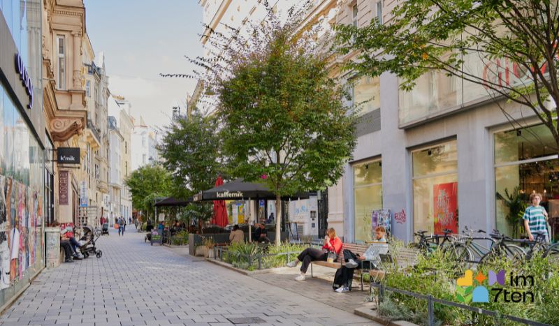 Menschen sitzen auf den Bänken in der Zollergasse, 1070 Wien, unter Bäumen und zwischen begrünten Beeten. © Eugenie Sophie Photography